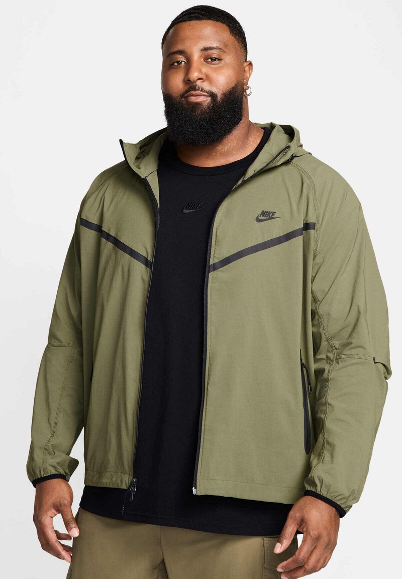 Khaki kurtka przeciwwiatrowa Nike z kapturem, czarnymi akcentami, kieszeniami na zamek i elastycznymi mankietami, nałożona na czarny T-shirt Nike.
