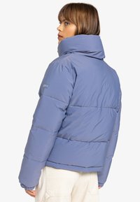 Dikke, blauwe damesjas met een hoge kraag, gewatteerd ontwerp en elastische manchetten. Logo op de bovenarm. Zachte, gladde textuur.