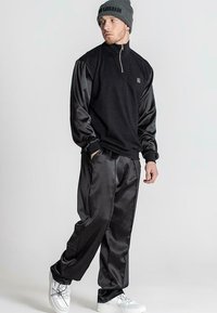 Černý fleecový pulovr s polovičním zipem a lesklými černými saténovými rukávy, spárovaný s volnými černými saténovými kalhotami a bílými teniskami.