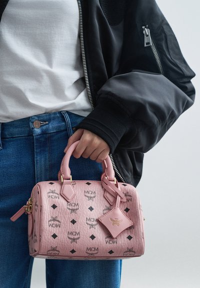 En person i blå jeans, vit skjorta och svart jacka håller en rosa MCM-handväska med svarta logotyper och guldfärgad detaljer.