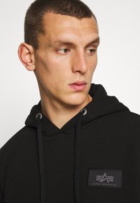 Sudadera negra hecha de una tela suave, con capucha ajustable y un parche de marca en el pecho. La capucha tiene ojales plateados.