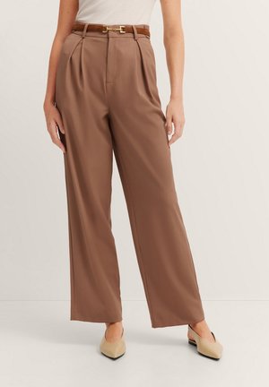 Braune hoch geschnittene Hose mit Falten, ausgestattet mit einem braunen Gürtel und einem leichten Glanz. Kombiniert mit beigefarbenen spitzen Schuhen.