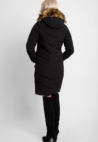 Mujer vestida con un abrigo acolchado negro hasta la rodilla con capucha de piel, y botas negras de tacón hasta la rodilla, de pie con las manos en los bolsillos.