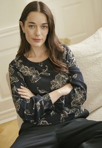 Donna con lunghi capelli castani, che indossa una blusa nera a fiori e pantaloni neri, seduta su un divano beige a trama con le braccia incrociate.