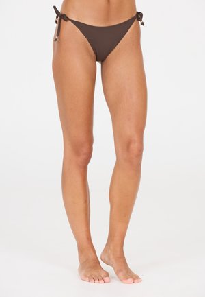 Jambes et pieds nus d'une personne portant un bas de bikini marron foncé avec des liens sur les côtés, debout devant un fond blanc uni.