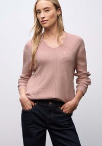 Vrouw met lang blond haar, gekleed in een roze ribbeltrui en donkere spijkerbroek, staand met haar handen in de zakken tegen een effen achtergrond.