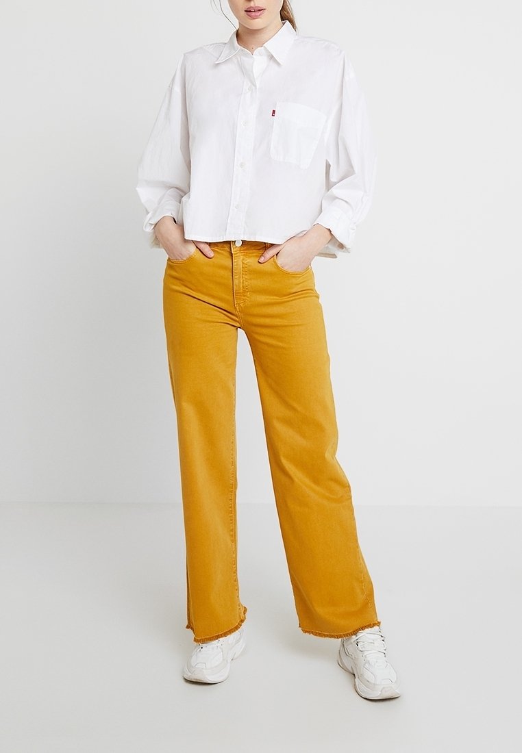 Camisa blanca recortada con botones y un bolsillo en el pecho, combinada con pantalones de tiro alto color mostaza con un dobladillo deshilachado y zapatillas blancas.