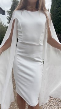 Žena v bielych priliehavých šatách s dlhými, voľne padajúcimi plášťovými rukávmi stojaca vonku na štrku s drevinami v pozadí.