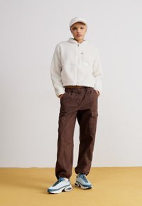 Fleece-Zip-Jacke in Weiß, braune Cargo-Hosen und mehrfarbige Sneakers. Das Model trägt eine weiße Mütze und steht vor einem gelben Hintergrund.