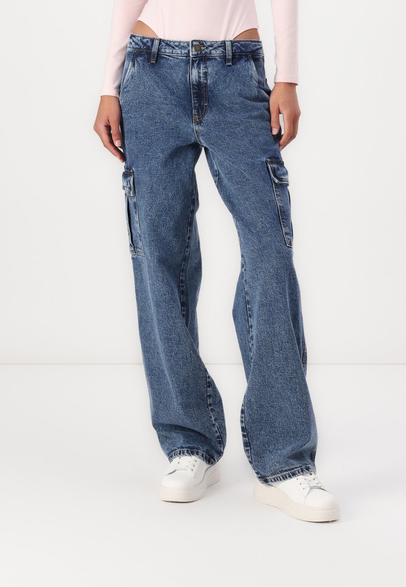 Modre denim cargo hlače z ohlapnim krojem, ki imajo stranske žepe in ravne noge. Obute z belimi športnimi copati.