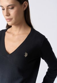 Maglione nero a v-neck realizzato in materiale morbido con bordi a coste e un piccolo logo dorato ricamato sul lato sinistro del petto.