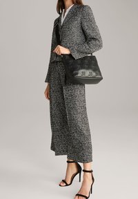 Borsa a spalla in pelle nera con motivo in rilievo, abbinata a un completo in tweed grigio composto da giacca e pantaloni a gamba larga.