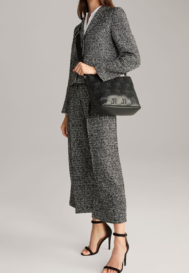 Borsa a spalla in pelle nera con motivo in rilievo, abbinata a un completo in tweed grigio composto da giacca e pantaloni a gamba larga.