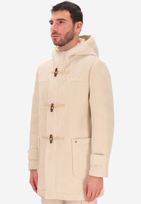 Uomo che indossa un cappotto in corduroy beige con cappuccio e tasche frontali a toppa, in piedi contro uno sfondo bianco uniforme.