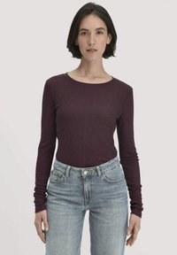 Frau mit kurzem, dunklem Haar, die ein enges, dunkelviolette Langarmoberteil und helle, hochtaillierte Jeans trägt, steht vor einem schlichten Hintergrund.