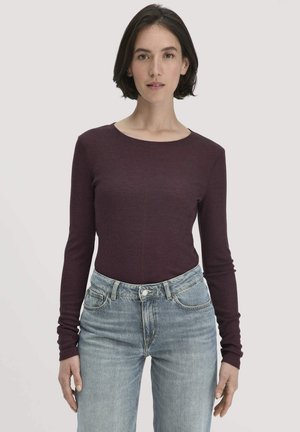 Vrouw met kort donker haar die een strakke donkerpaarse lange mouwtop en lichtblauwe high-waisted jeans draagt, staande tegen een effen achtergrond.