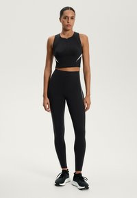 Fekete sportcrop top, elülső cipzárral, testhezálló fekete leggingsel, fehér oldalsávokkal párosítva. Fekete sportcipővel viselve.