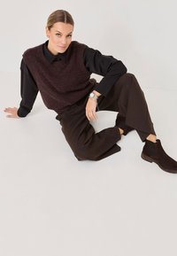 Brązowa dzianinowa kamizelka na czarną koszulę z kołnierzykiem, w połączeniu z szerokimi brązowymi spodniami i ciemnymi botkami za kostkę. Prosty srebrny zegarek na nadgarstku.