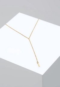 Collier délicat en or en forme de Y avec une barre horizontale et un pendentif en barre verticale suspendue, présenté sur une surface blanche inclinée.