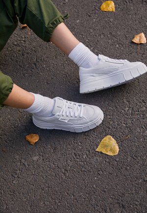 Jambes portant des baskets blanches Puma et des chaussettes blanches sur un bitume avec des feuilles d’automne jaunes éparpillées.