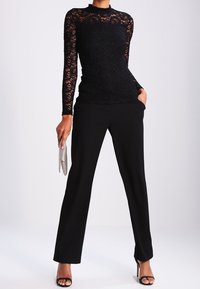 Sort blonde langærmet top med høj hals, parret med sorte lige bukser. Modellen holder en sølv clutch. Sorte højhælede sandaler med åben tå.