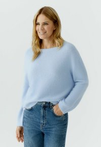 Lächelnde Frau mit blondem Haar, die einen hellblauen, flauschigen Pullover und blaue Jeans trägt, mit einer Hand in der Tasche, vor einfarbig hellem Hintergrund.