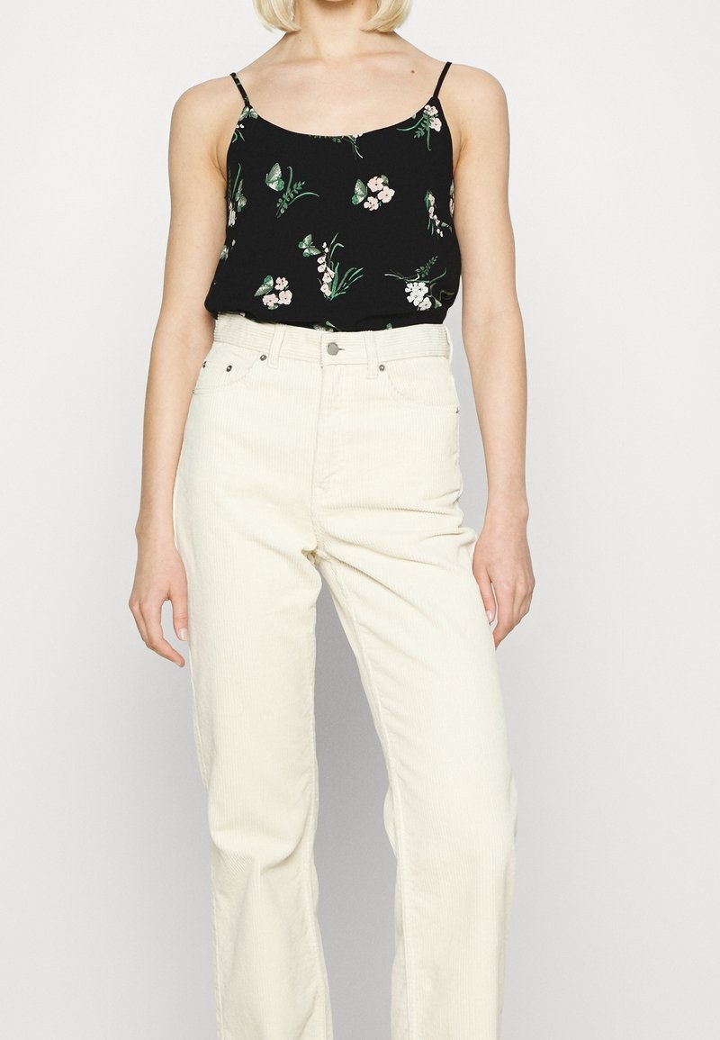 Camisola nera a fiori con spalline sottili abbinata a pantaloni larghi in corduroy color crema a vita alta, con un focus sui dettagli di texture e pattern.