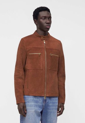 Homme portant une veste en daim marron avec une fermeture éclair dorée et des détails de poche, associée à un jean bleu clair, debout devant un fond clair uni.
