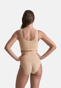Donna con capelli lunghi, che indossa un'intimo beige a vita alta e un top corto abbinato, in piedi di spalle con le mani sui fianchi.