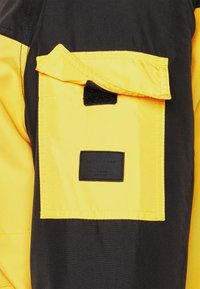 Czarna i żółta kurtka z wyraźną żółtą kieszonką, zamykaną na czarną taśmę rzep. Na kieszeni znajduje się czarna metka z napisem "JACK&JONES."