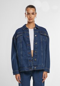 Donna con i capelli raccolti indossa una giacca di jeans blu scuro oversize su una maglietta bianca corta e pantaloni in denim coordinati, su uno sfondo semplice.