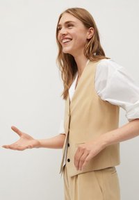 Un gilet sans manches beige sur une chemise blanche. Il présente un design ajusté avec des boutons noirs, des poches sur les côtés et une texture lisse.