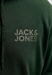 Σκούρο πράσινο ύφασμα με γκρι κείμενο "JACK & JONES" και πράσινο κορδόνι ορατό στην αριστερή πλευρά.