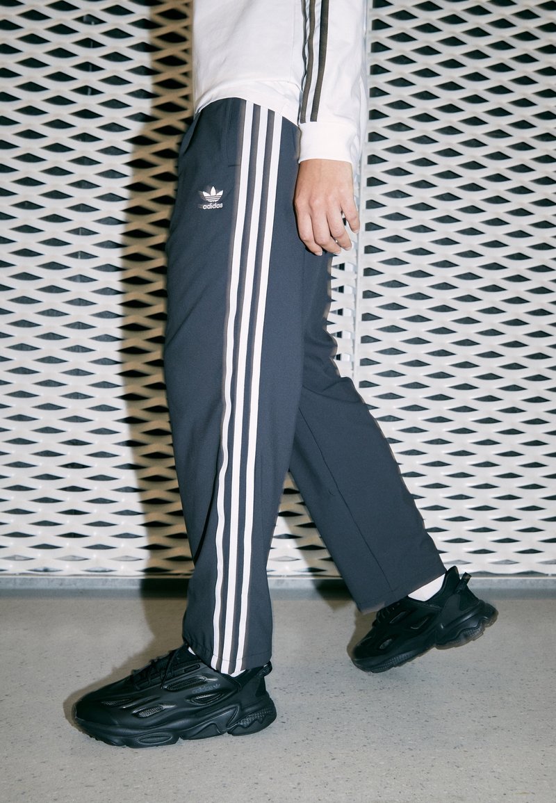Gray athletic pants with three white stripes along the side, featuring an embroidered logo. Paired with black sneakers, textured surface.