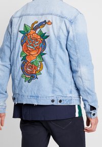 Ljusblå jeansjacka med en stor broderad design av en orm som slingrar sig runt röda och orange rosor med gröna blad.