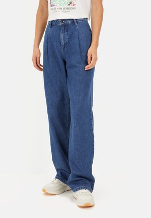 Hoge taille, rechte jeans van middelblauwe denim. Heeft plooien aan de voorkant en een gestikte zoom. Draagt met een wit graphic t-shirt.