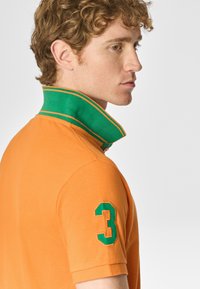 Polo arancione con colletto verde a due righe e il numero 3 ricamato in verde sulla manica sinistra. Tessuto dalla superficie liscia.