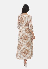 Robe longue et fluide à motifs floraux en crème avec des motifs marron. Elle possède des manches évasées et une taille cintrée, avec un détail de jupe plissée. Des talons transparents sont portés.