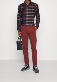 Man som står mot en neutral bakgrund, iförd en rutig skjorta och röda byxor, håller en liten svart väska och bär sportskor.