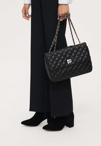 Black quilted handbag with a silver clasp, featuring a chain strap. Paired with black wide-leg pants and pointed-toe heeled boots.