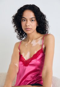 Camisole en satin de couleur rose vif avec un délicat bord en dentelle rouge, dotée de fines bretelles et d'une texture lisse.