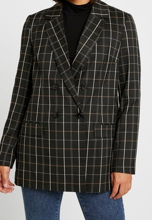 Blazer noir croisé avec un motif quadrillé blanc et marron porté sur un col roulé noir et un jean bleu.