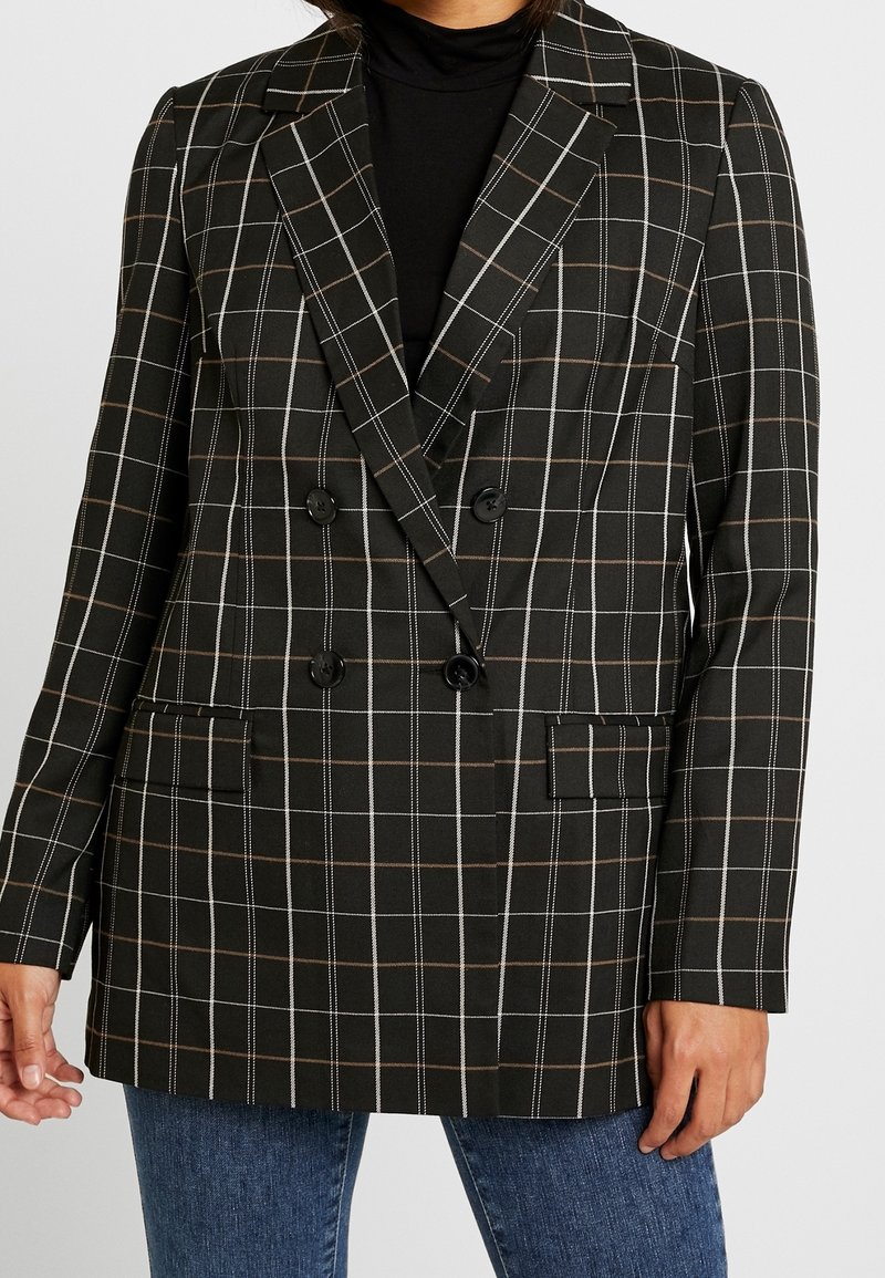 Blazer noir croisé avec un motif quadrillé blanc et marron porté sur un col roulé noir et un jean bleu.