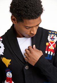 Giovane che guarda in basso, indossando un cardigan nero con disegni festivi che includono un soldatino, una campana, un pupazzo di neve e Babbo Natale.