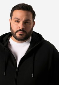 Černá mikina na zip s měkkou texturou, s kapucí na šňůrku a bílým vnitřním tričkem. Materiál vypadá pohodlně a ležérně.