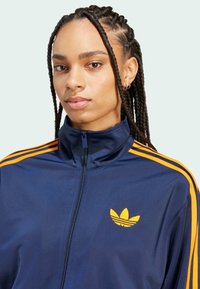 Dunkelblaue Zip-Up-Trainingsjacke mit gelbem Drei-Streifen-Design auf den Schultern und besticktem Logo auf der Brust. Glatte Textur, sportlicher Schnitt.