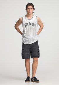 Canottiera bianca senza maniche con righe nere e stampa "SAN DIEGO", abbinata a pantaloni cargo neri con multiple tasche. Sneakers nere.