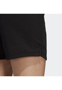 Gros plan sur un short noir sur la cuisse d'une personne, présentant une petite fente latérale et des détails de couture visibles.