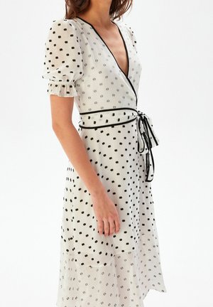 Femme portant une robe portefeuille blanche à pois noirs, manches bouffantes, bordure noire et ceinture nouée à la taille, vue de l'épaule au mollet.