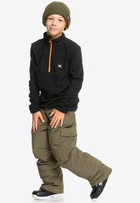 Pull en polaire noir avec fermeture éclair orange, pantalon cargo vert olive, bottes de neige noires et bonnet en tricot vert olive. Pas de motifs visibles.
